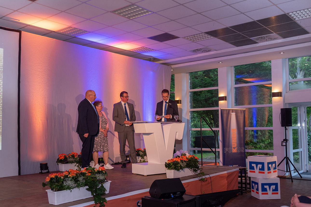 Auf der Bühne am Rednerpult steht Stefan Korte. Im Hintergrund Eduard Kolkmann und seine Frau sowie Norbert Scholtholt.