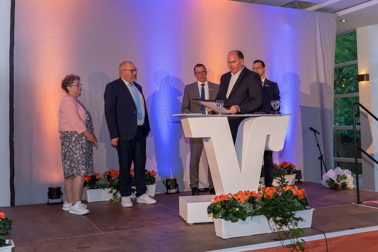 Carsten Waldhelm liest eine Laudatio auf der Bühne am Rednerpult vor. Im Hintergrund stehen Eduard Kolkmann mit seiner Frau und die Vorstände Stefan Korte und Norbert Scholtholt.