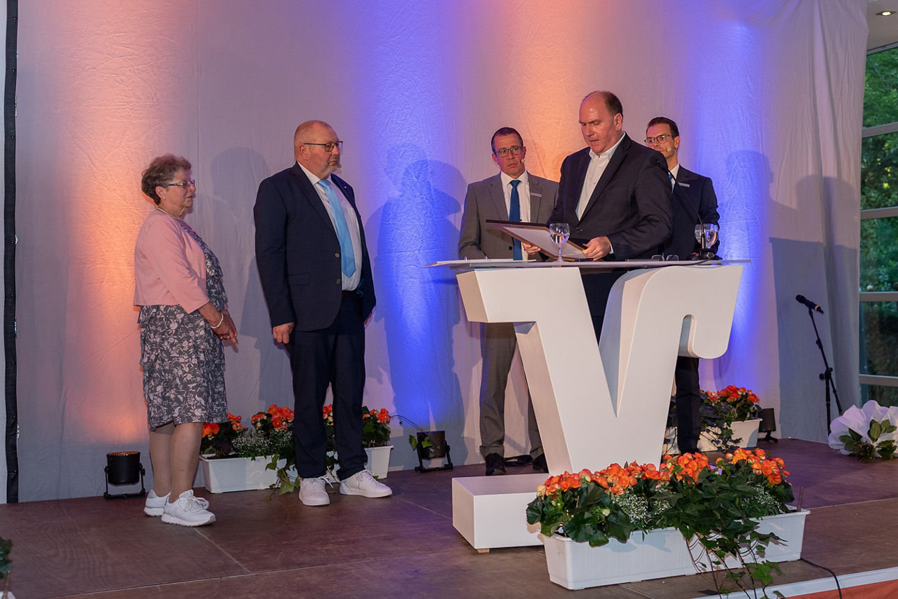 Carsten Waldhelm liest eine Laudatio auf der Bühne am Rednerpult vor. Im Hintergrund stehen Eduard Kolkmann mit seiner Frau und die Vorstände Stefan Korte und Norbert Scholtholt.