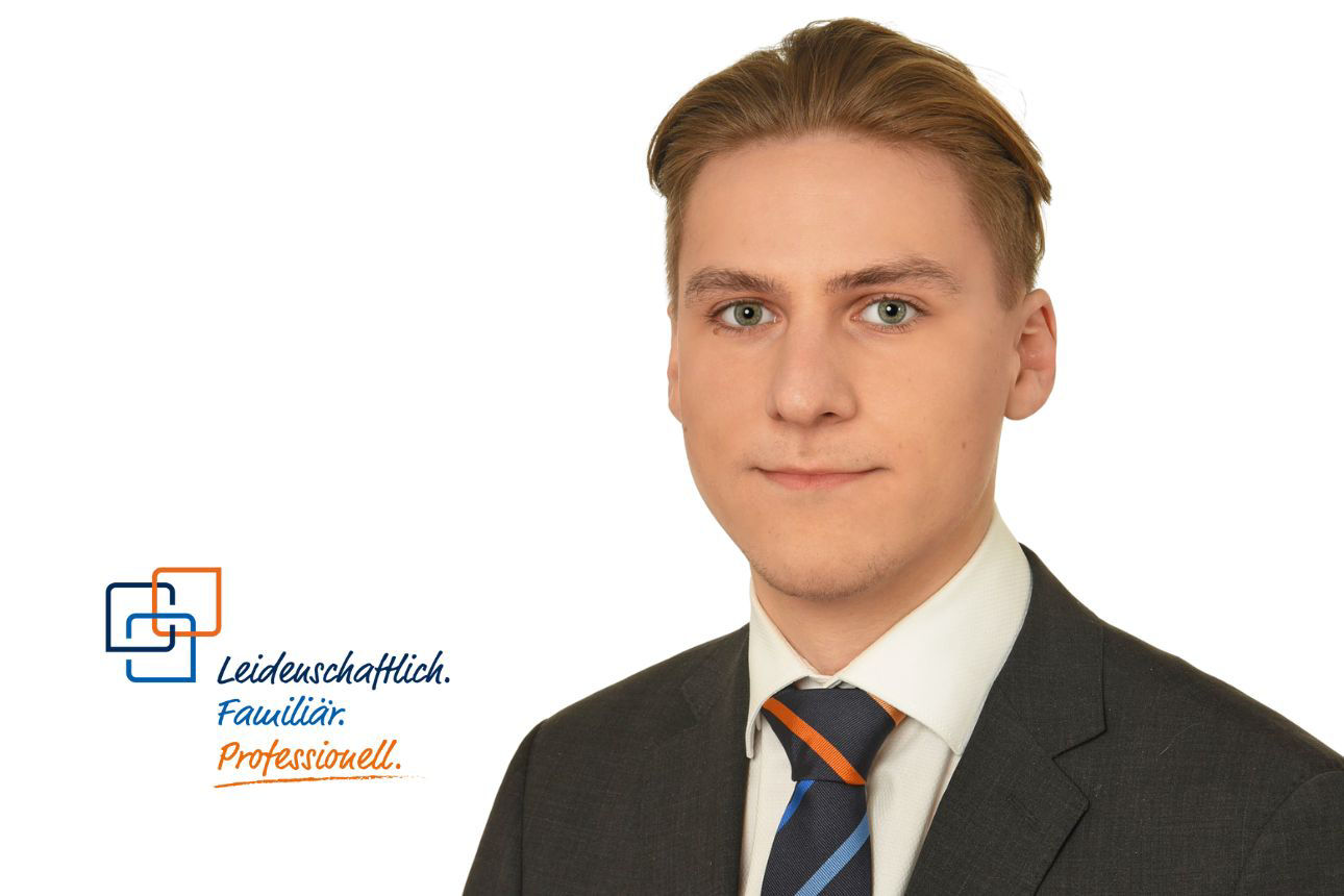 Valentin Brückner, ein junger Mann mit kurz geschnittenen blonden Haaren, trägt einen dunklen Anzug mit einer gestreiften blau-orangenen Krawatte und blickt ernst in die Kamera.