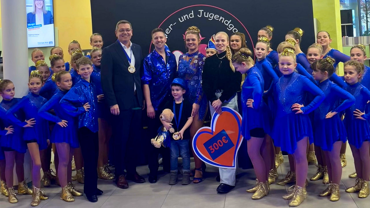 Kindergarde in blauen Kostümen steht aufgereiht der Größe nach vor grauer Wand mit Logo des Vereins. Mittig das Prinzenpaar umrahmt von Bankvorstand B.Nickel und Bankmitarbeiterin A.Gula
