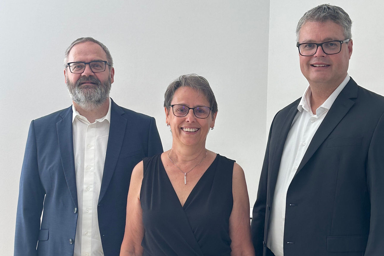Es stehen der Bereichsleiter Rechnungswesen, die Jubilarin und Vorstandsmitglied Markus Feser im Bankgebäude nebeneinander. Sie lächeln in die Kamera. 