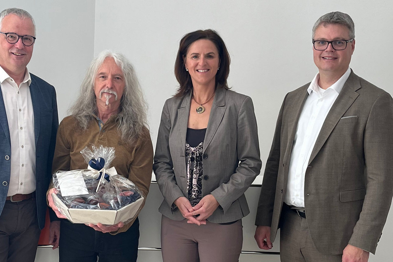 Auf dem Bild stehen drei Männer und eine Frau.  Einer der Männer hält einen Geschenkkorb in der Hand. Das Bild wurde in einem Bankgebäude aufgenommen. 