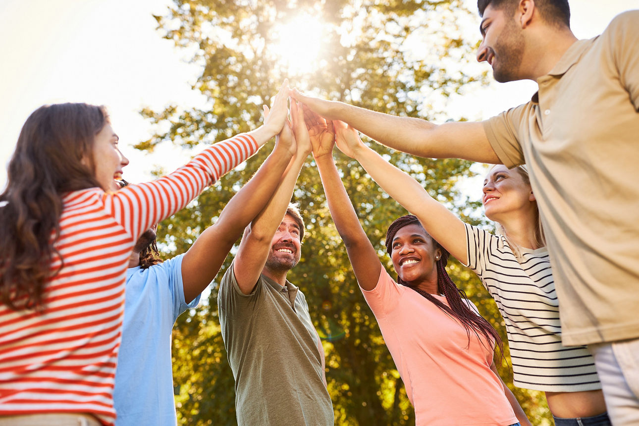 Gruppe von fünf Personen steht im Kreis und hebt die Hände zusammen nach oben, im Hintergrund Bäume und Sonne.
