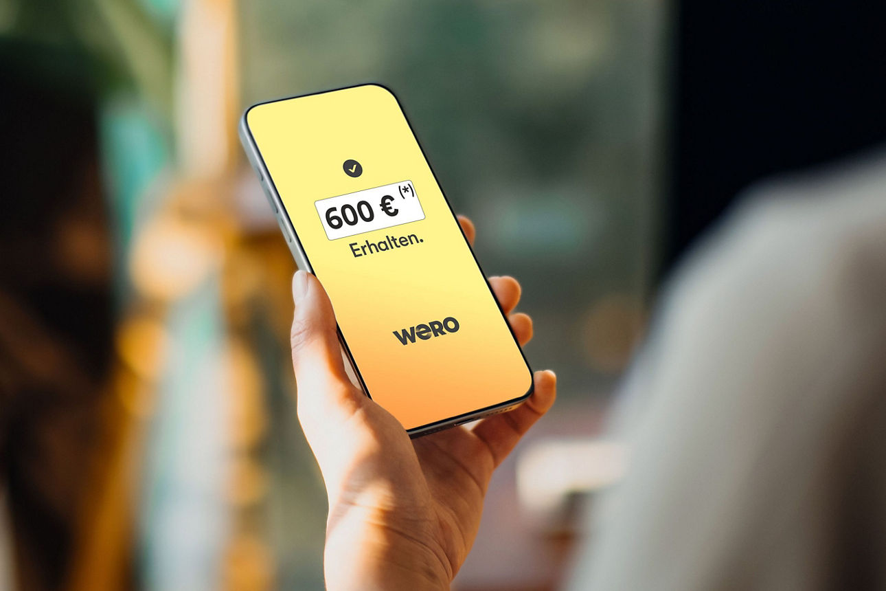 Eine Hand hält ein Smartphone mit gelbem Display und dem Text „600 €* Erfüllen. weco“.