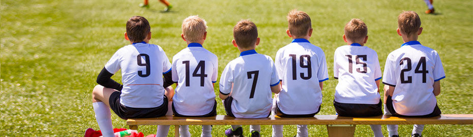 Sechs Jungen in weißen Fußballtrikots sitzen auf einer Holzbank auf einer Wiese
