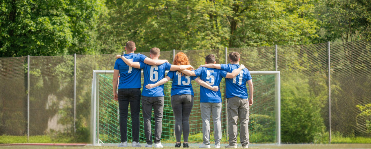 Fünf Personen stehen in blauen Trikots mit weißen Nummern auf dem Rücken vor einem Fußballtor. Sie haben die Arme umeinander 