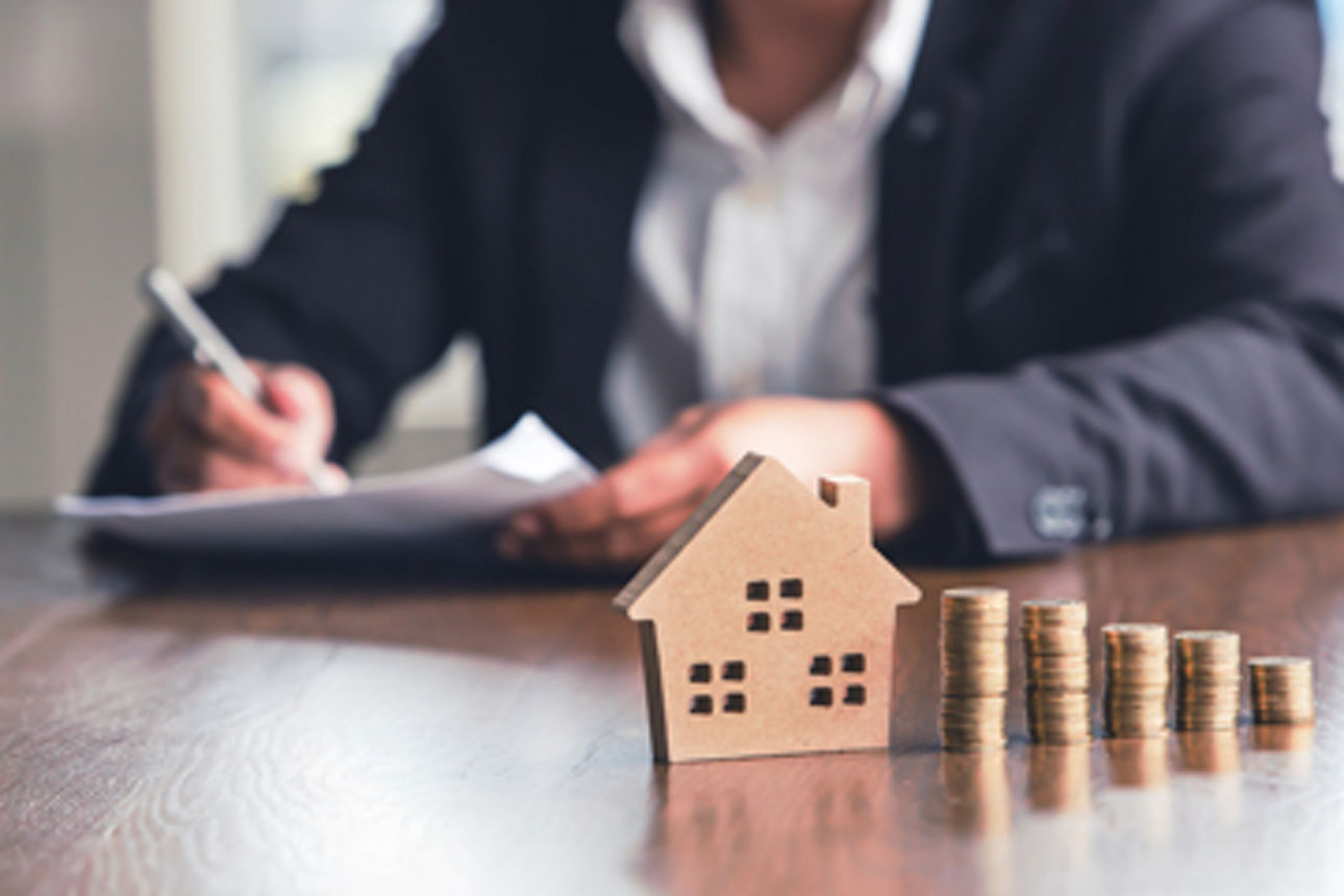 Ein Mann sitzt an einem Tisch und hält Dokumente in der Hand. Im Fokus ist ein Hausmodell mit absteigenden Münzstapeln.