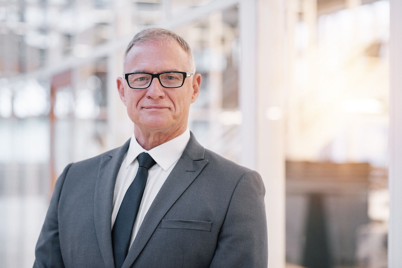 Ein älterer Mann mit Brille, grauem Anzug und Krawatte schaut freundlich.