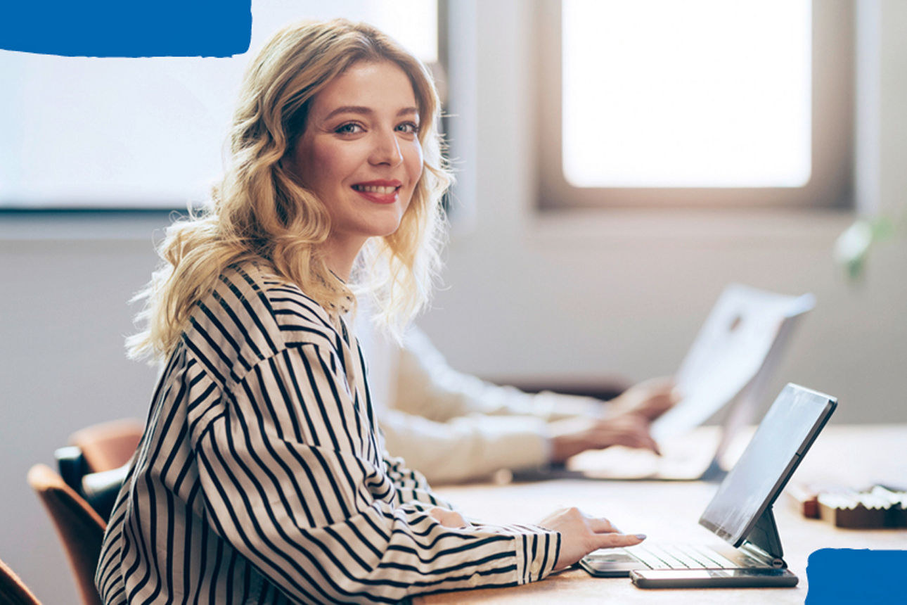 Junge Frau sitzt am Schreibtisch am Tablet und schaut in die Kamera