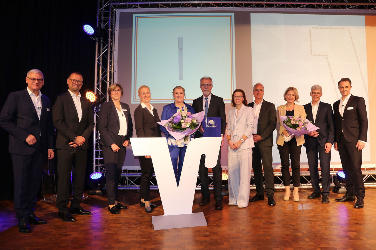 Das Bild zeigt ein Gruppenfoto auf der Bühne. In der Mitte steht der scheidende Aufsichtsratsvorsitzende Roland Denecke.