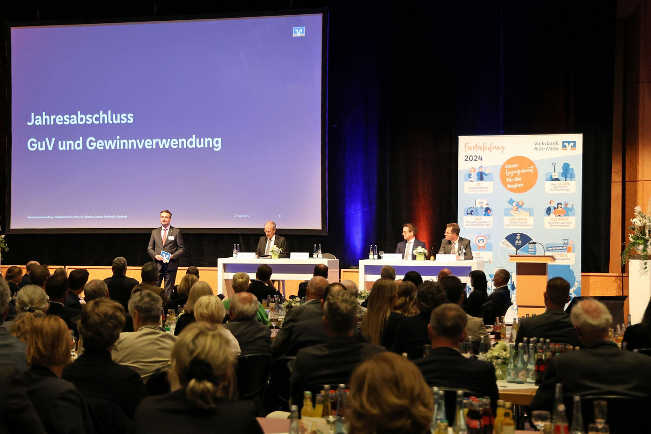 Auf der Bühne der Vertreterversammlung steht eine männliche Person in Anzug und Krawatte, drei weitere Personen sitzen an hohen Tischen. Von links nach rechts: Jörg Lott und Ingo Abrahams aus dem Vorstand der Volksbank Ruhr Mitte, des weiteren Wernher Schwarz und Stephan Dieler aus dem Aufsichtsrat.