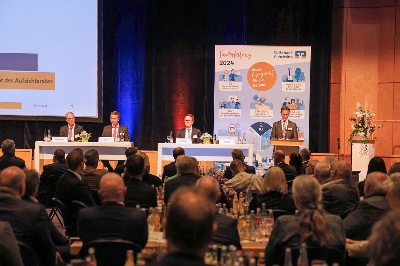 Man sieht auf einer kleinen Bühne zwei Hochtische, an denen insgesamt 3 Personen sitzen. Neben den Tischen ist ein Rednerpult, an dem ein Mann steht. Im HIntergrund sieht man den Teil einer Leinwand, einen schweren Bühenvorhang und ein großes Roll-up.Im Vordergrund des Bildes sieht man Besucher der Veranstaltung von hinten, wie sie auf die Bühne schauen. Das Bild wurde aufgenommen auf der Vertreterversammlung der Volksbank Ruhr Mitte in 2025.