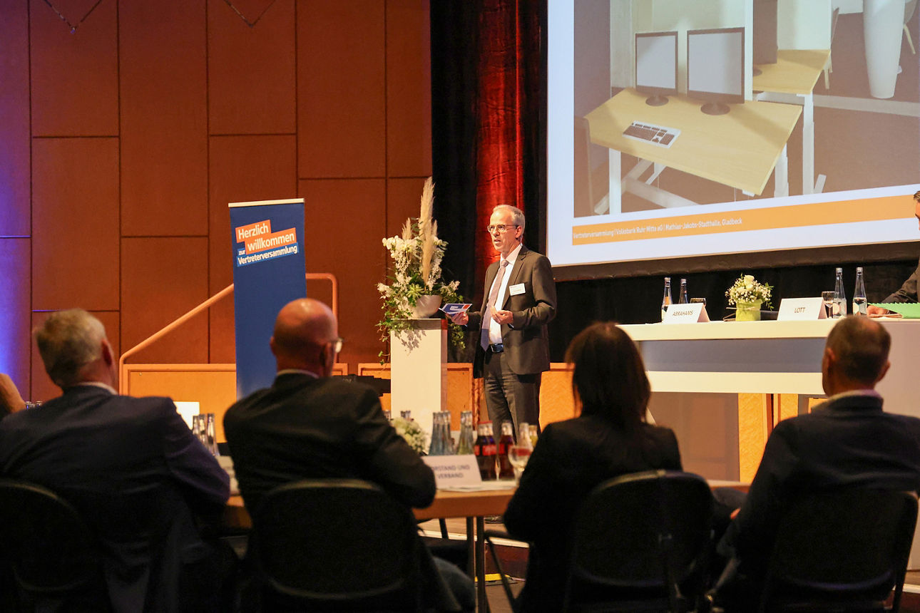 Man sieht auf einer kleinen Bühne unseren Vorstand Ingo Abrahams, wie er zum Publikum.spricht.Im HIntergrund sieht man den Teil einer Leinwand.Im Vordergrund des Bildes sieht man Besucher der Veranstaltung von hinten, wie sie auf die Bühne schauen. Das Bild wurde aufgenommen auf der Vertreterversammlung der Volksbank Ruhr Mitte in 2025.