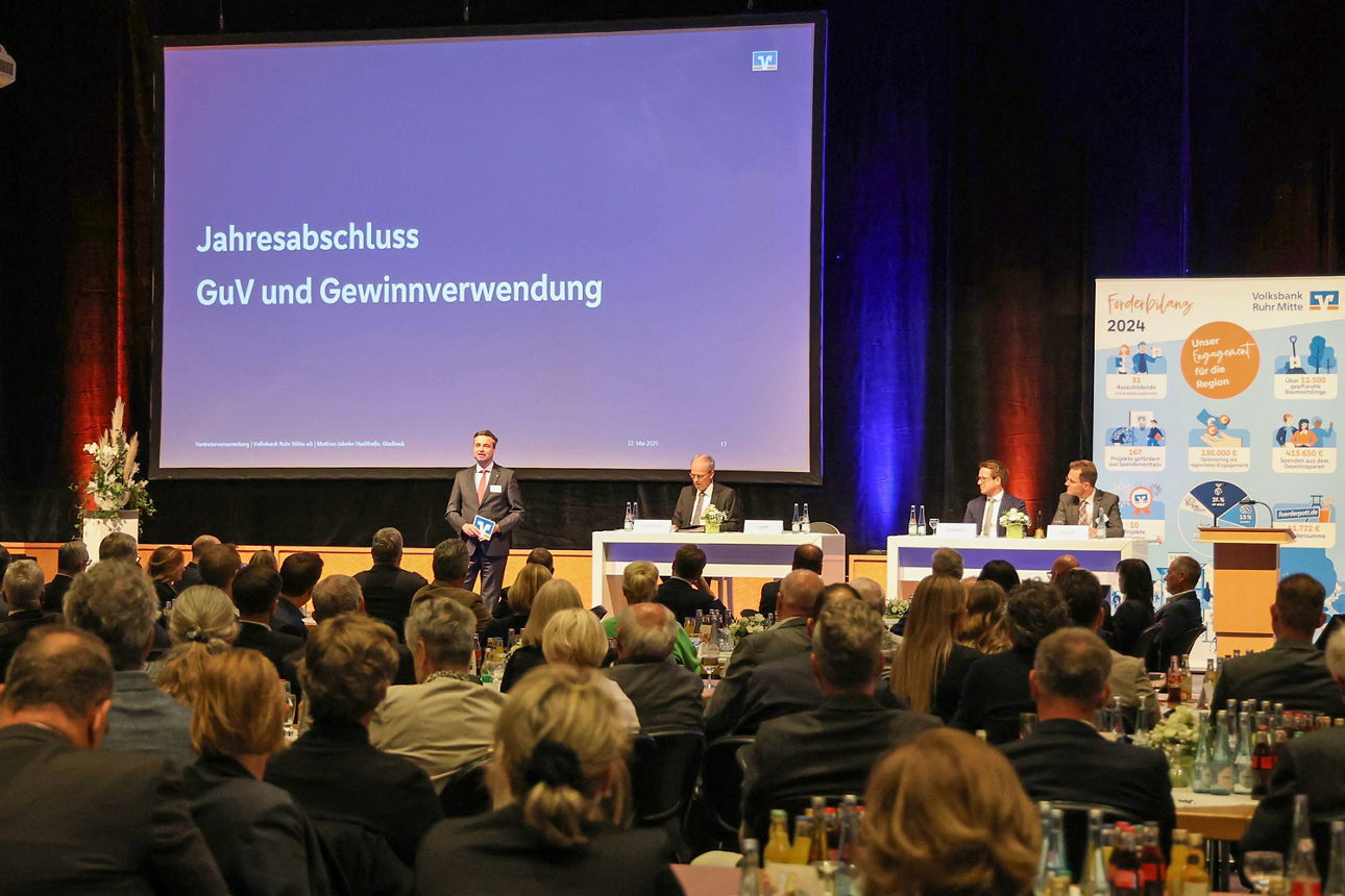 Man sieht auf einer kleinen Bühne zwei Hochtische, an denen insgesamt 3 Personen sitzen. Neben den Tischen ist ein Rednerpult, an dem keiner steht. Links neben den Tischen steht unser Vorstand Jörg Lott und spricht zum Publikum.Im HIntergrund sieht man eine Leinwand, einen schweren Bühenvorhang und ein großes Roll-up.Im Vordergrund des Bildes sieht man Besucher der Veranstaltung von hinten, wie sie auf die Bühne schauen. Das Bild wurde aufgenommen auf der Vertreterversammlung der Volksbank Ruhr Mitte in 2025.