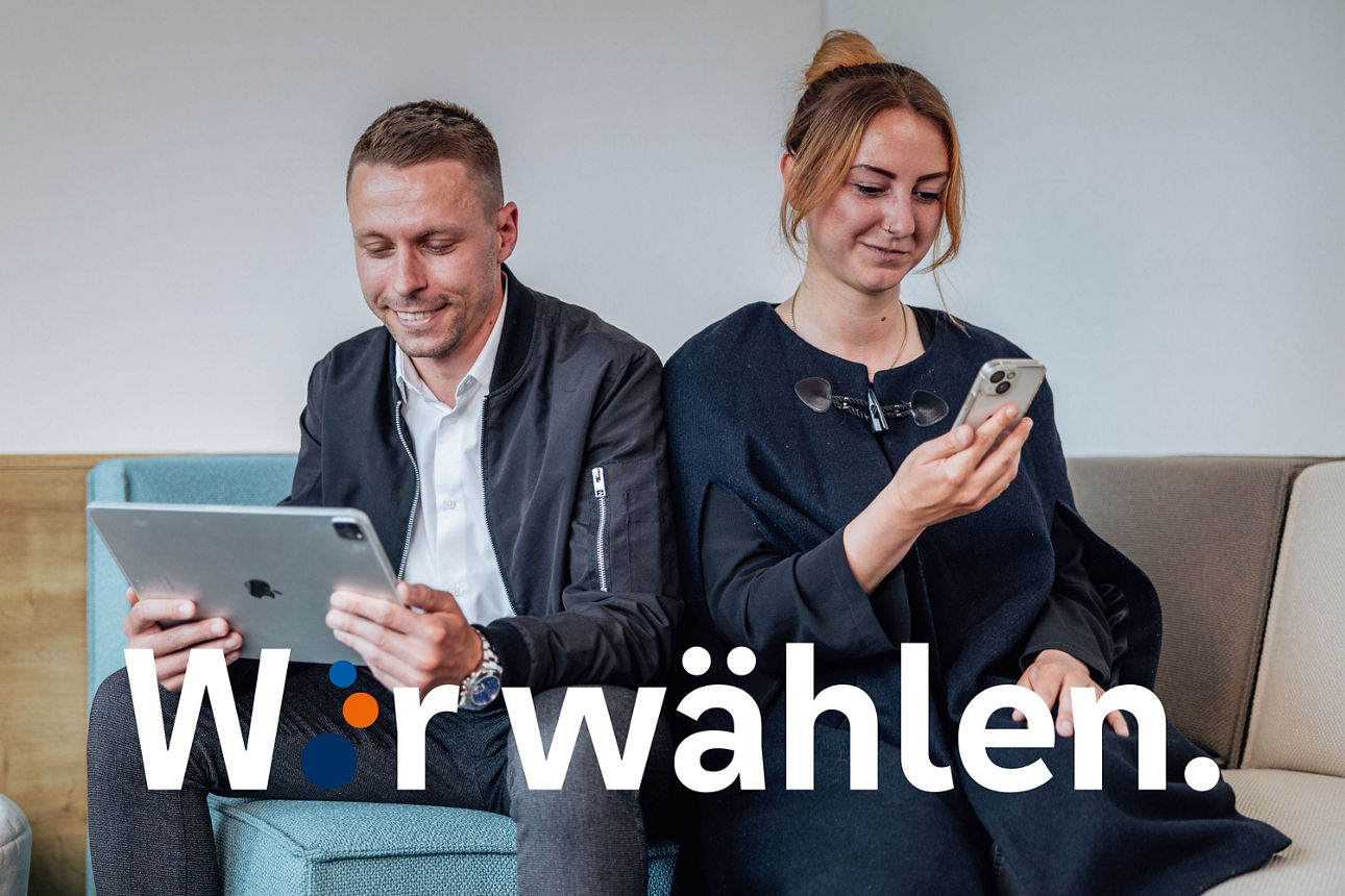 Ein junger Mann sitzt mit dem Tablet auf der Couch und lächelt es an. Eine junge Frau sitzt direkt daneben und hat ein Handy in der Hand und lächelt auch hinein. 