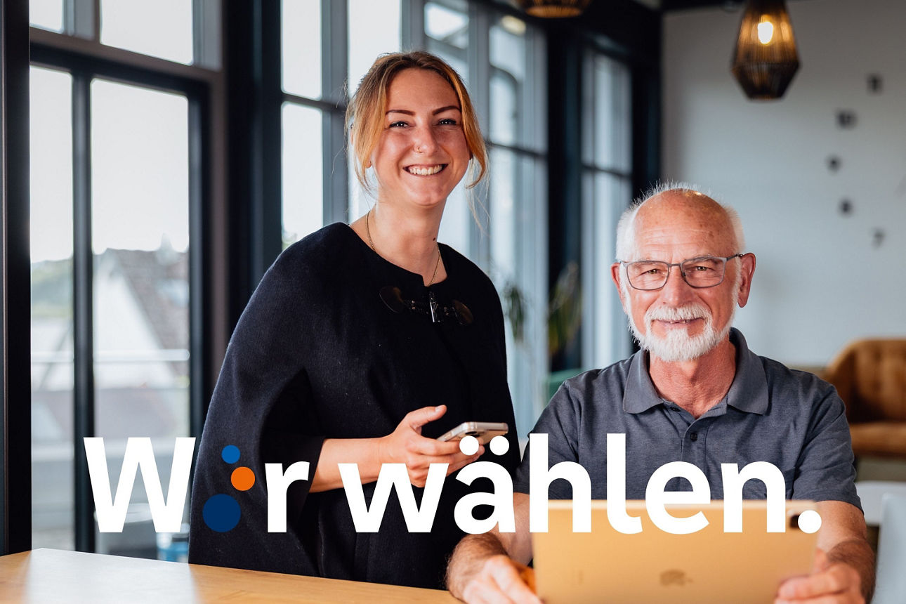 Eine junge Frau steht an einem hohen Tisch, lächelt in die Kamer und hält dabei ihr Handy in der Hand. Ein älterer Mann sitzt daneben, hält ein Tablet vor sich und lächelt ebenfalls in die Kamera. 
