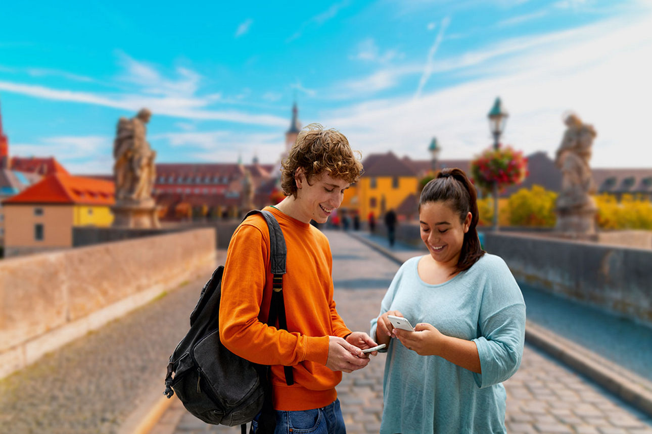 Zwei junge Menschen stehen auf der Alten Mainbrücke in Würzburg und schauen lächelnd auf ihre Handys