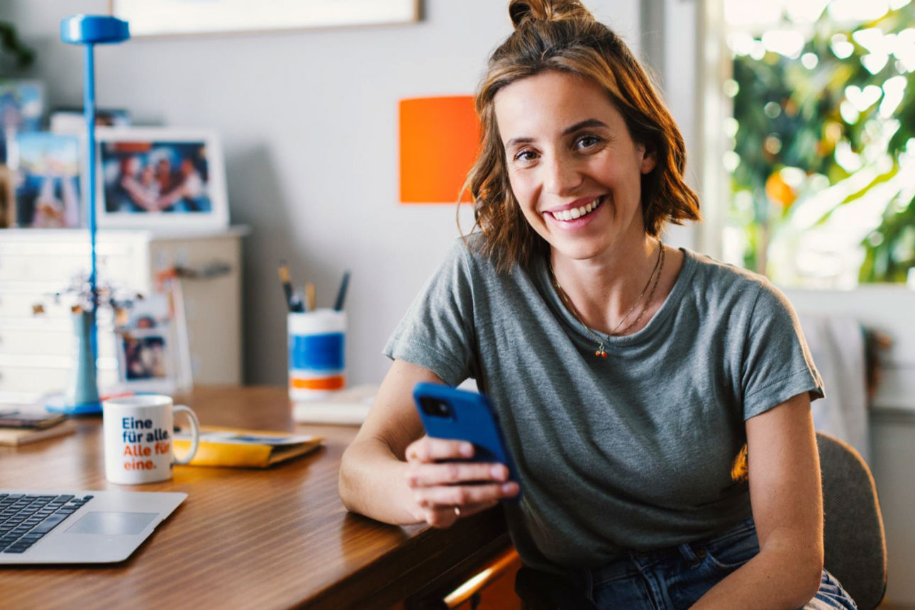 Junge Frau am Schreibtisch mit Handy in der Hand.