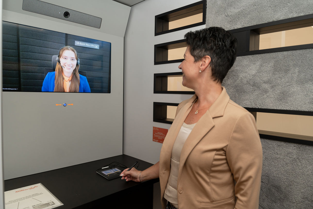 Frau steht in Videokabine und unterhält sich mit Mitarbeiterin über das Display
