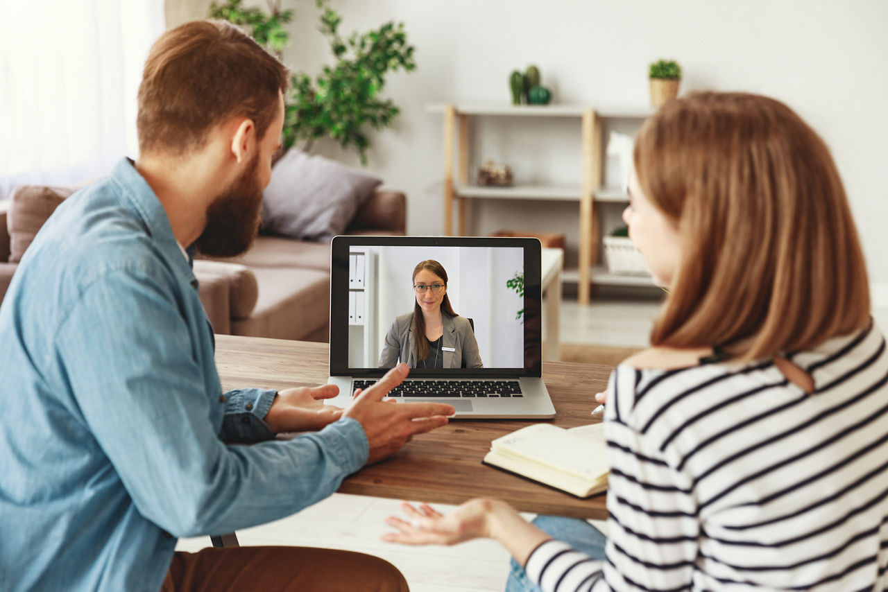 Mann und Frau sitzen im Wohnzimmer vor einem Laptop und sprechen mit ihrer Beraterin