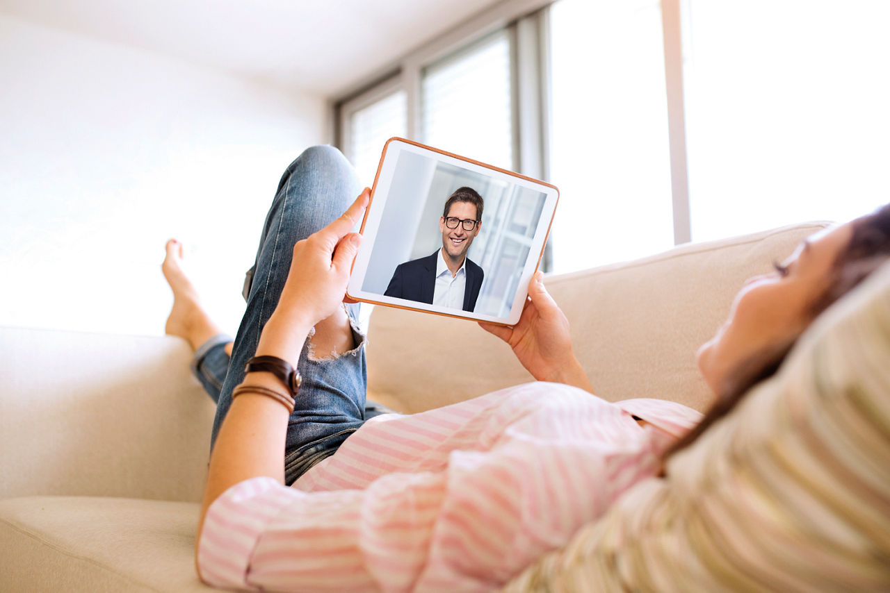 Abbildung zeigt eine Frau, die mit ihrem Tablet auf der Couch liegt und gerade eine Videoberatung mit Meine Bank durchführt