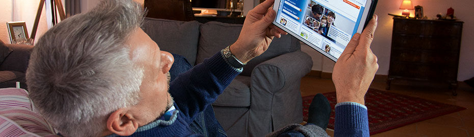 Ein älterer Mann liegt auf dem Sofa und hält ein Tablet in der Hand. Auf dem Bildschirm ist eine Website mit Informationen und Bildern zu sehen.