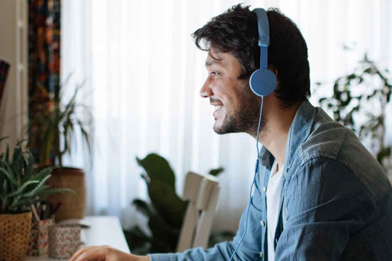 Ein Mann sitzt lächelnd am Schreibtisch, trägt Kopfhörer und arbeitet konzentriert an einem Computer in einer gemütlichen Woh