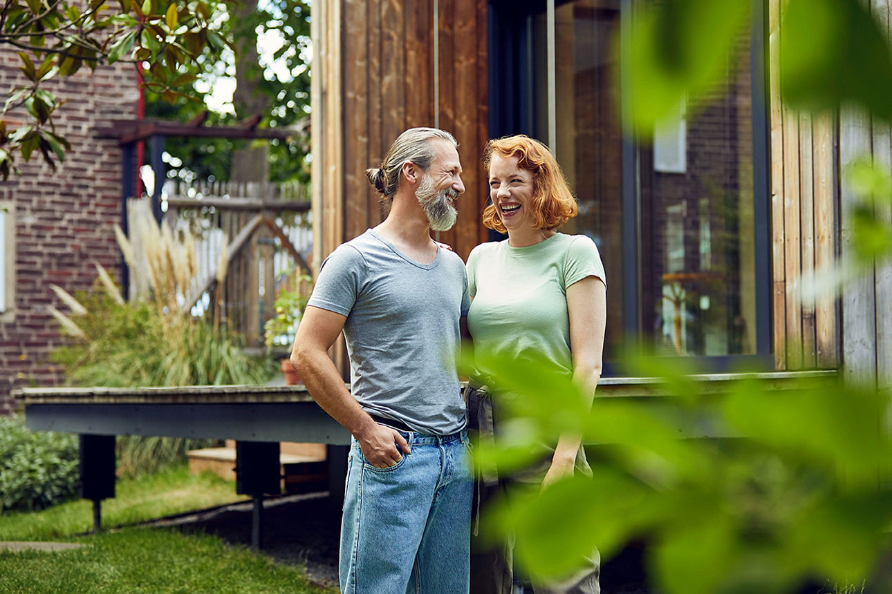 Fröhliches, europäisch gelesenes, Paar steht in Deutschland, in ihrem Garten vor dem eigenen, modernen Tinyhouse. Mann und Frau sind zwischen 30-40 Jahren alt. Die Szene steht für Erfolg im Leben und Green living.