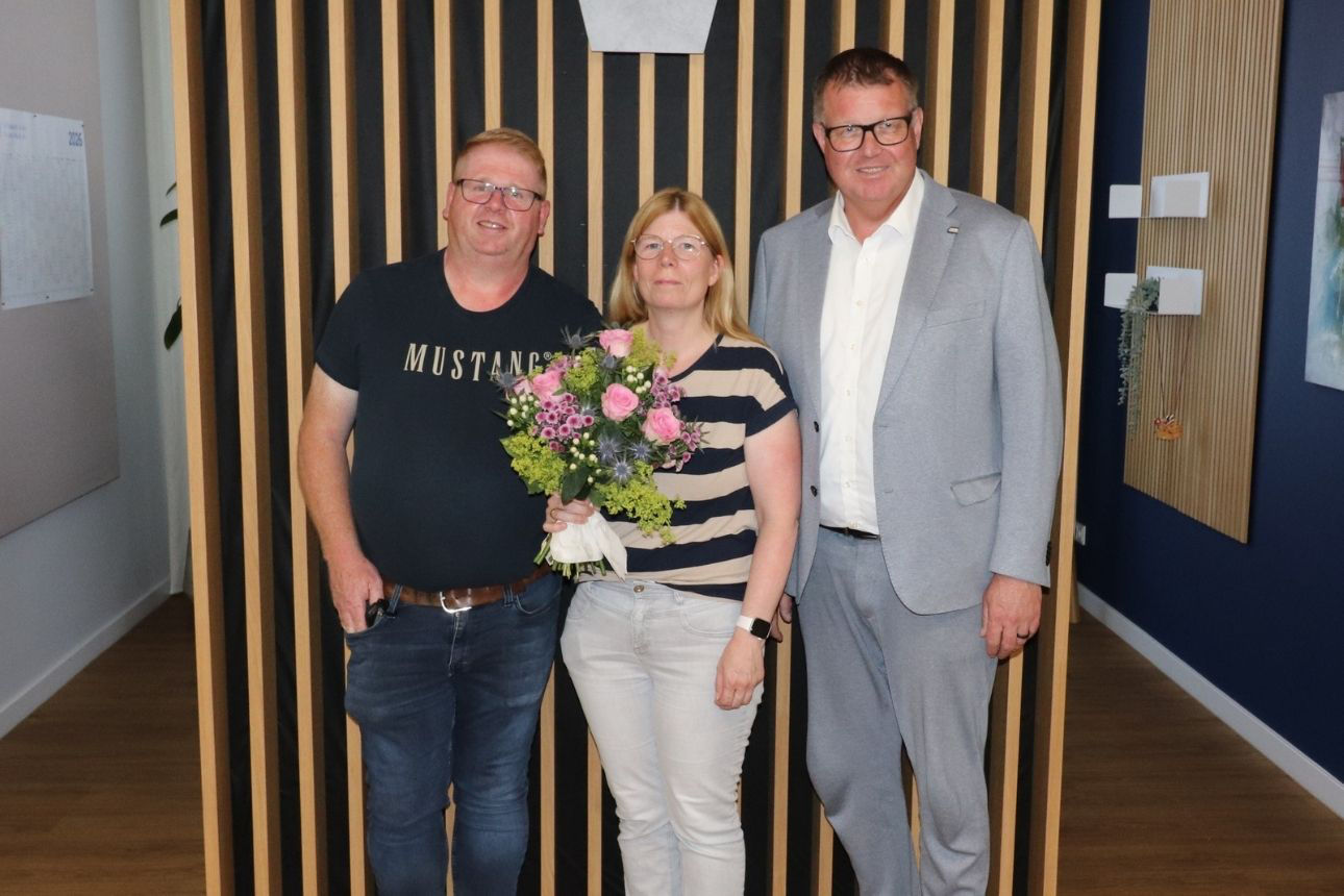 Drei Personen stehen in den Räumlichkeiten einer Bank vor einer Holzlamellenwand; die Person in der Mitte hält einen Blumenstrauß, die beiden anderen Personen stehen daneben.