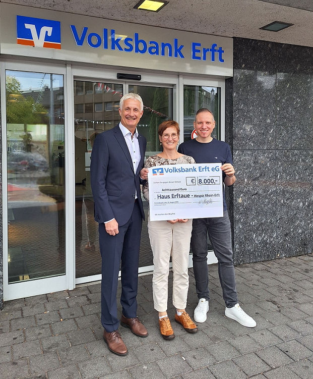 Zwei Herren und eine Dame halten einen Spendenscheck vor einer Bankfiliale hoch.