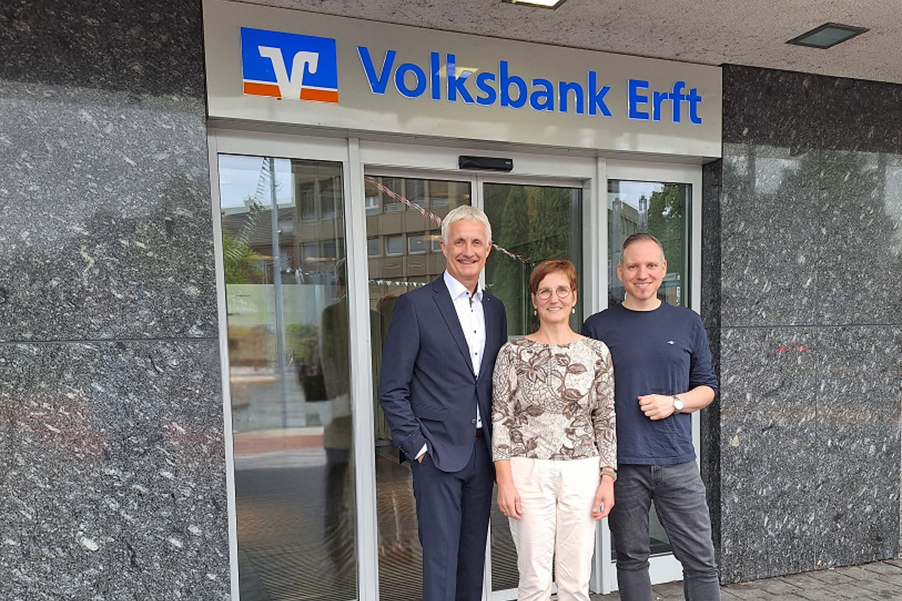 Zwei Herren und eine Dame stehen vor einer Bankfiliale und lächeln in eine Kamera.