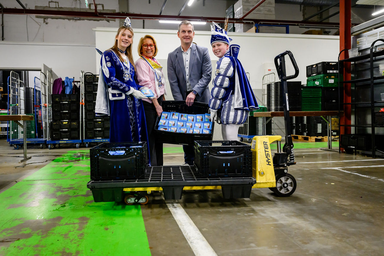 Auf dem Foto stehen vier Personen in einer Lagerhalle der Tafel. Zwei Kinder tragen blaue Karnevalskostüme.