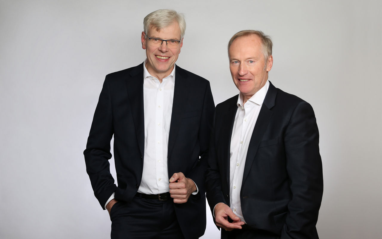 Zwei Männer in dunklen Anzügen stehen zusammen vor einem schlichten weißen Hintergrund und strahlen Professionalität und Sel