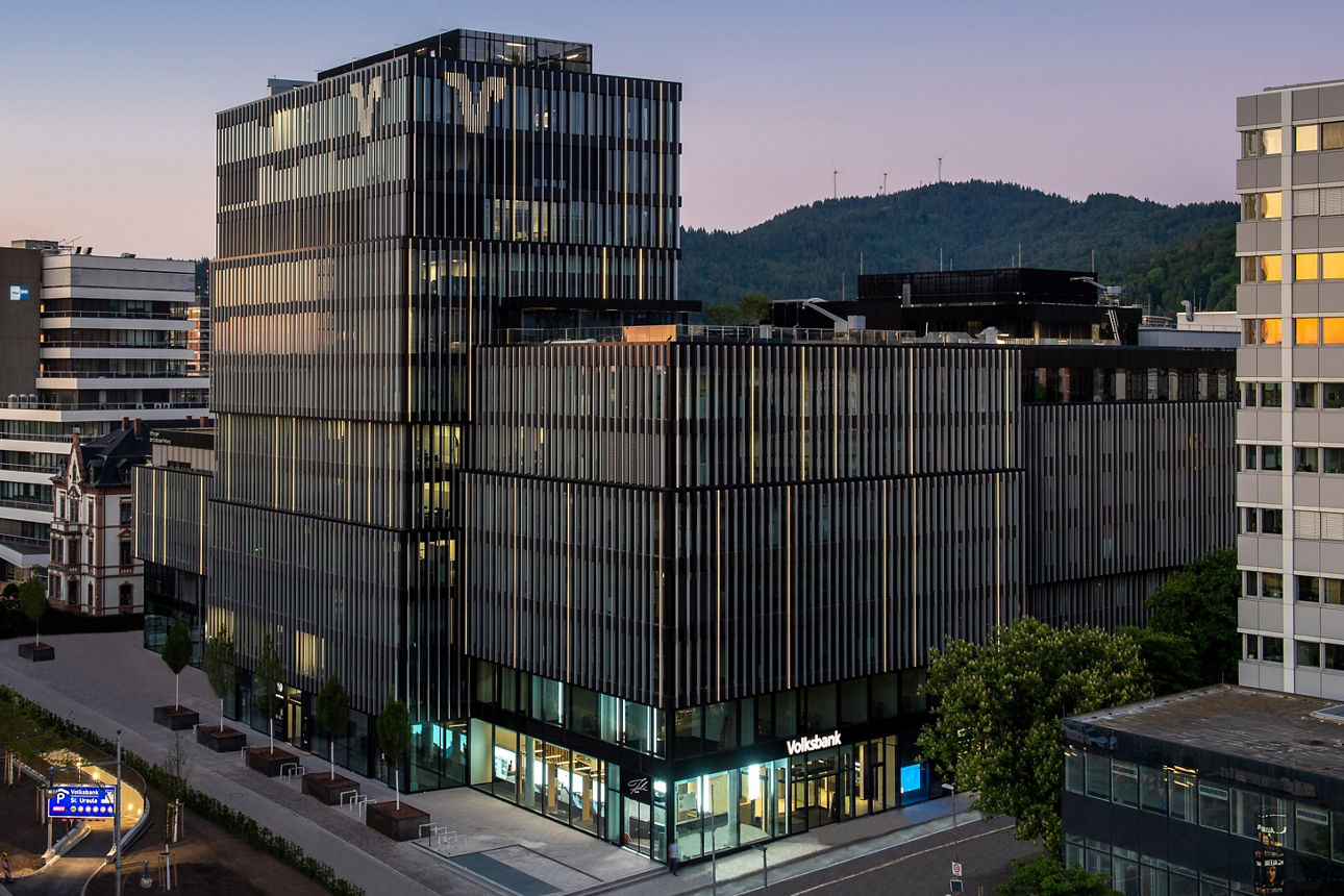 Modernes Bürogebäude der Volksbank Freiburg mit Glasfassaden, im Hintergrund bewaldete Hügel bei Sonnenuntergang