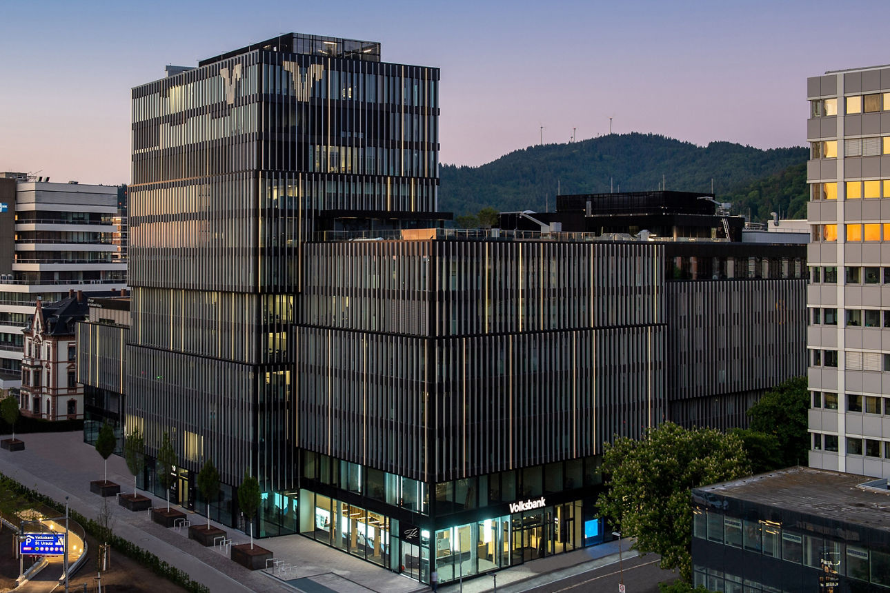 Modernes Bürogebäude der Volksbank Freiburg mit Glasfassaden, im Hintergrund bewaldete Hügel bei Sonnenuntergang