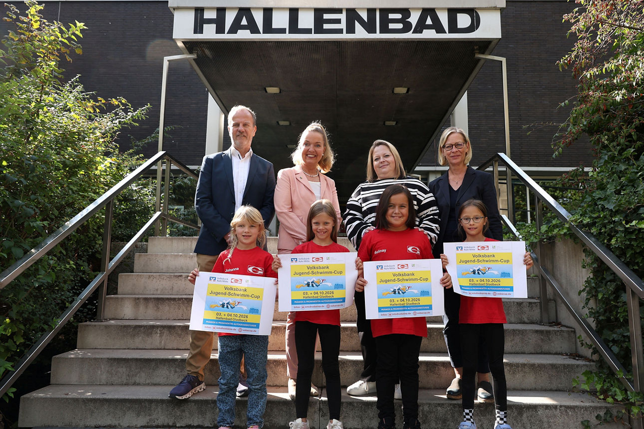 Gruppenfoto vor dem Eingang des Hallenbads: Vier Kinder halten Plakate zum „Volksbank Jugend-Schwimm-Cup“ in den Händen, hinter ihnen stehen vier Erwachsene, darunter die Bürgermeisterin, auf den Treppenstufen. Die Gruppe posiert vor dem Gebäude, das von Grünflächen und Geländern eingerahmt wird.