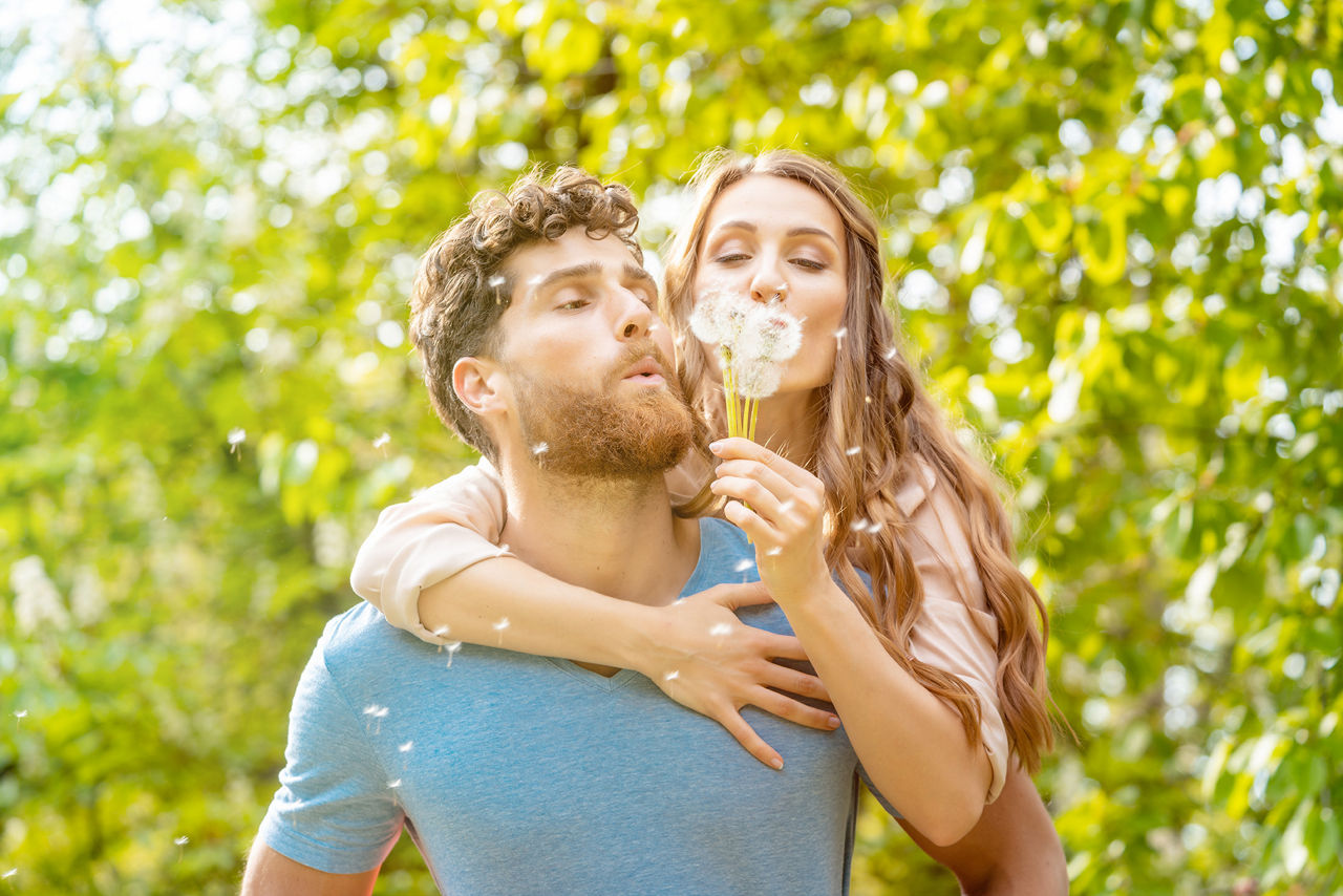 Eine Frau und ein Mann in der Natur. Er trägt sie auf dem Rücken und sie pusten eine Pusteblume