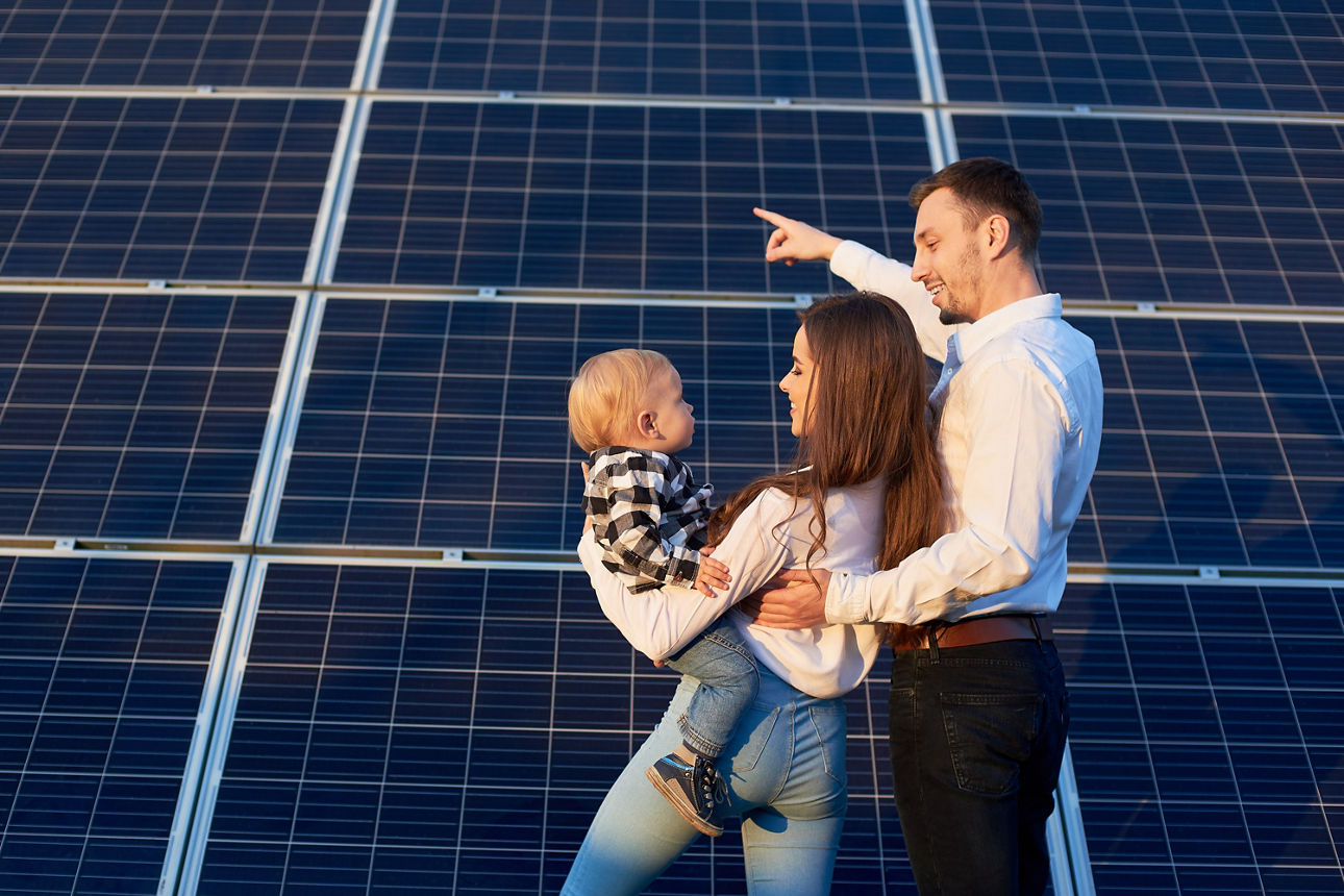 Rückansicht einer jungen, modernen Familie, die gemeinsam mit ihrem Baby an einer Photovoltaikanlage steht – sonniger Tag