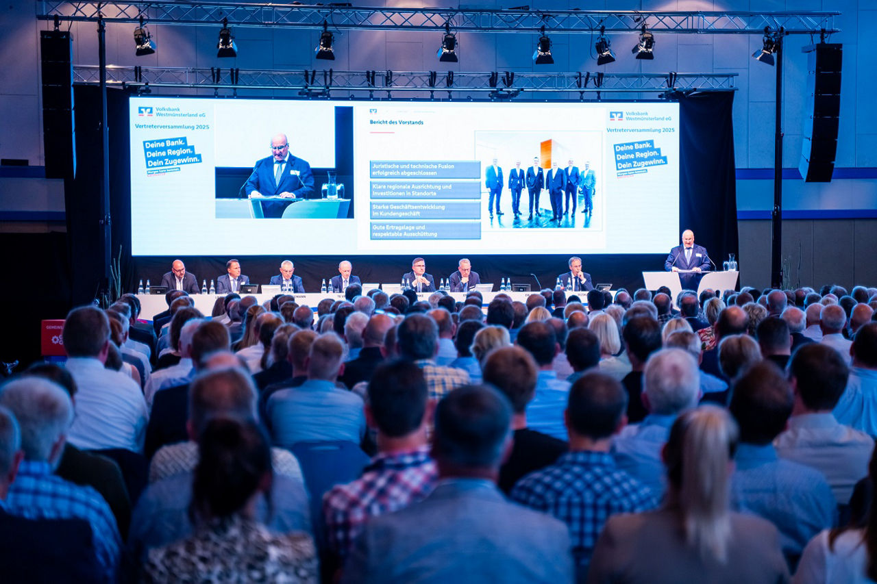 Viele Menschen sitzen vor einer Bühne auf der der Vorstand der Volksbank Westmümsterland sitzt 