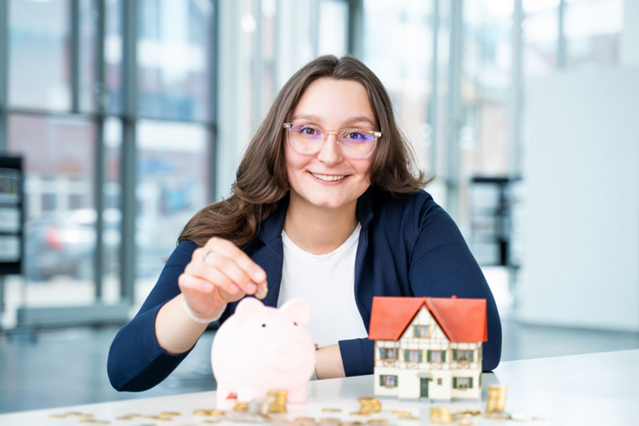 Frau sitzt vor einem Tisch auf dem Münzen liegen  und ein Haus steht 
