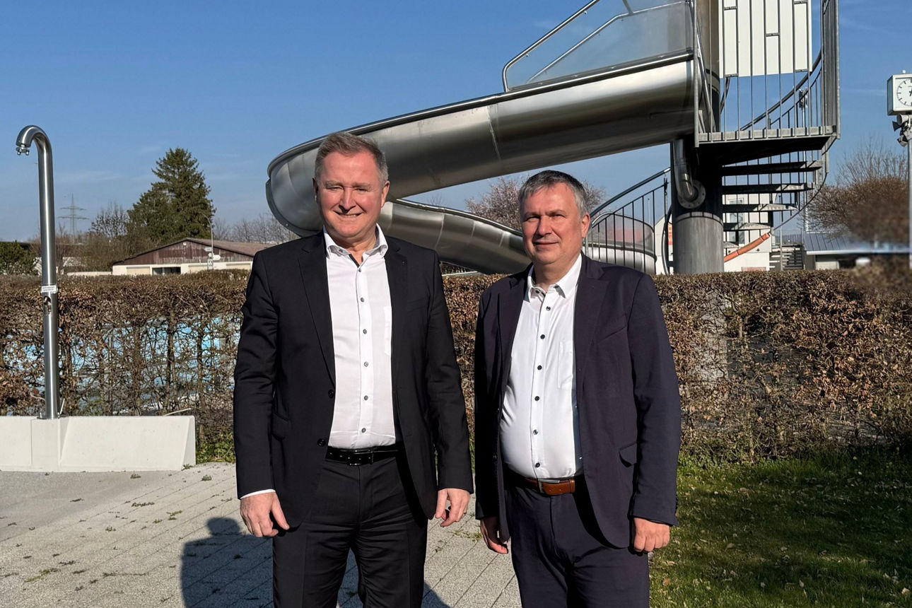 Die zwei Bankvorstände stehen im Freibad vor einer großen Wasserrutsche.