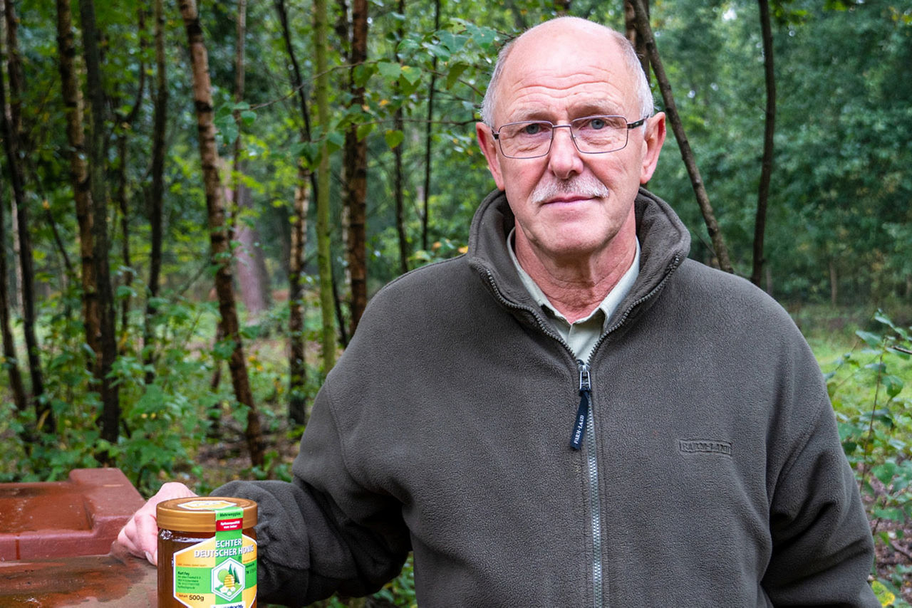 Imker steht an Bienenstock mit Honigglas in der Hand