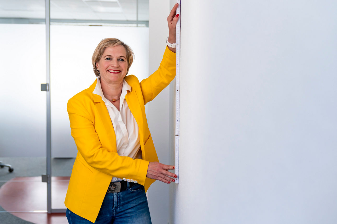 Frau steht lächelnd mit einem Zollstock an der Wand uns misst was aus.
