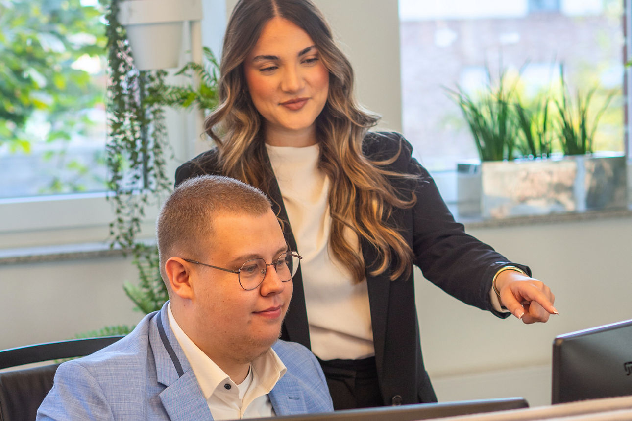 An Mann sitzt an einem Schreibtisch, eine junge Frau steht hinter ihm und zeigt auf seinen Monitor