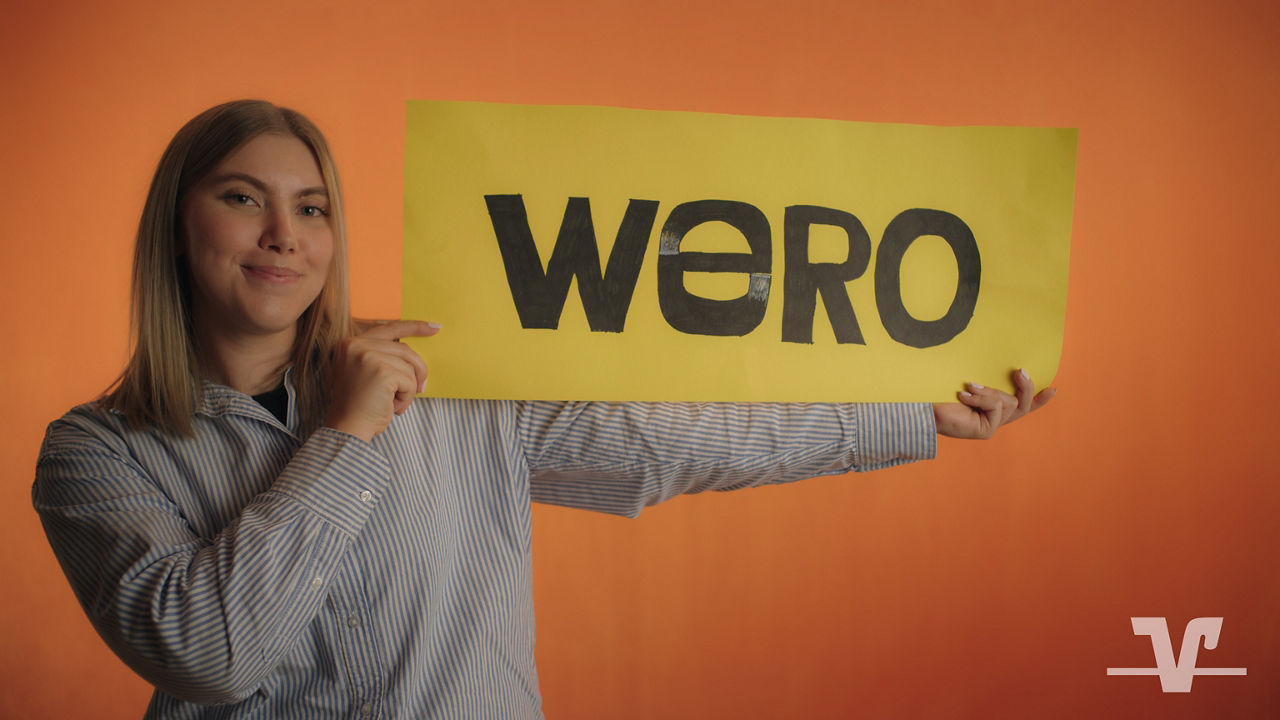 Person im gestreiften Hemd hält gelbes Plakat mit „wero“ vor orangefarbenem Hintergrund