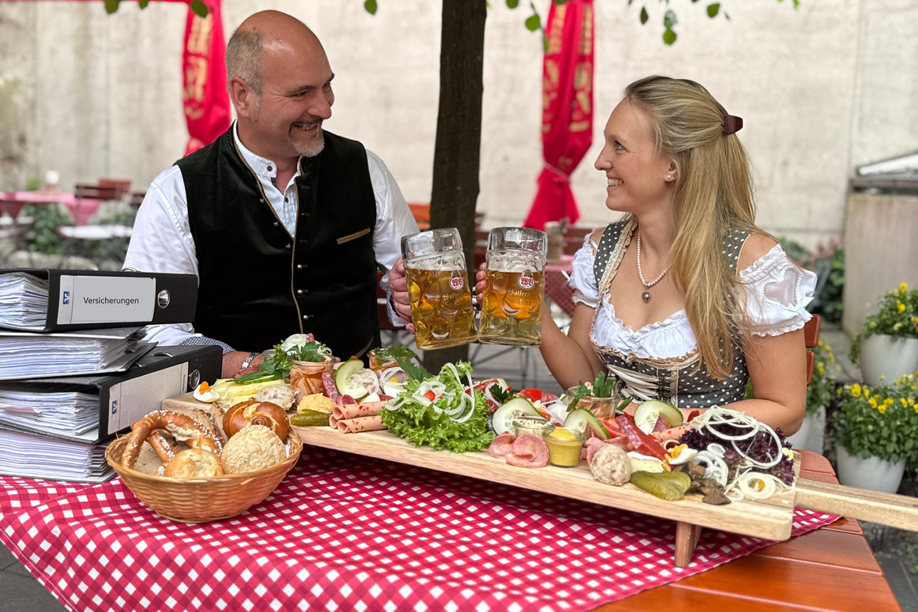 2 Personen stoßen mit Bierkrug an und sitzen in Biergarten hinter Tisch mit Versicherungsordnern und Brotzeit