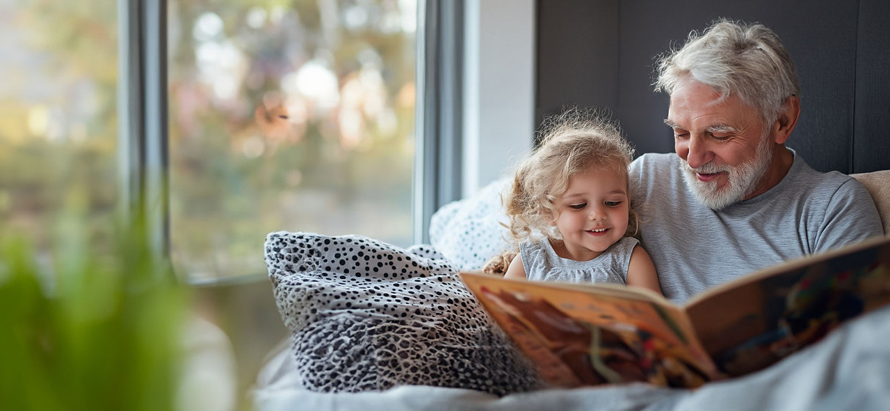 Älterer Mann sitzt mit kleinem Mädchen gemeinsam auf einem Sofa und schauen in ein Kinderbuch
