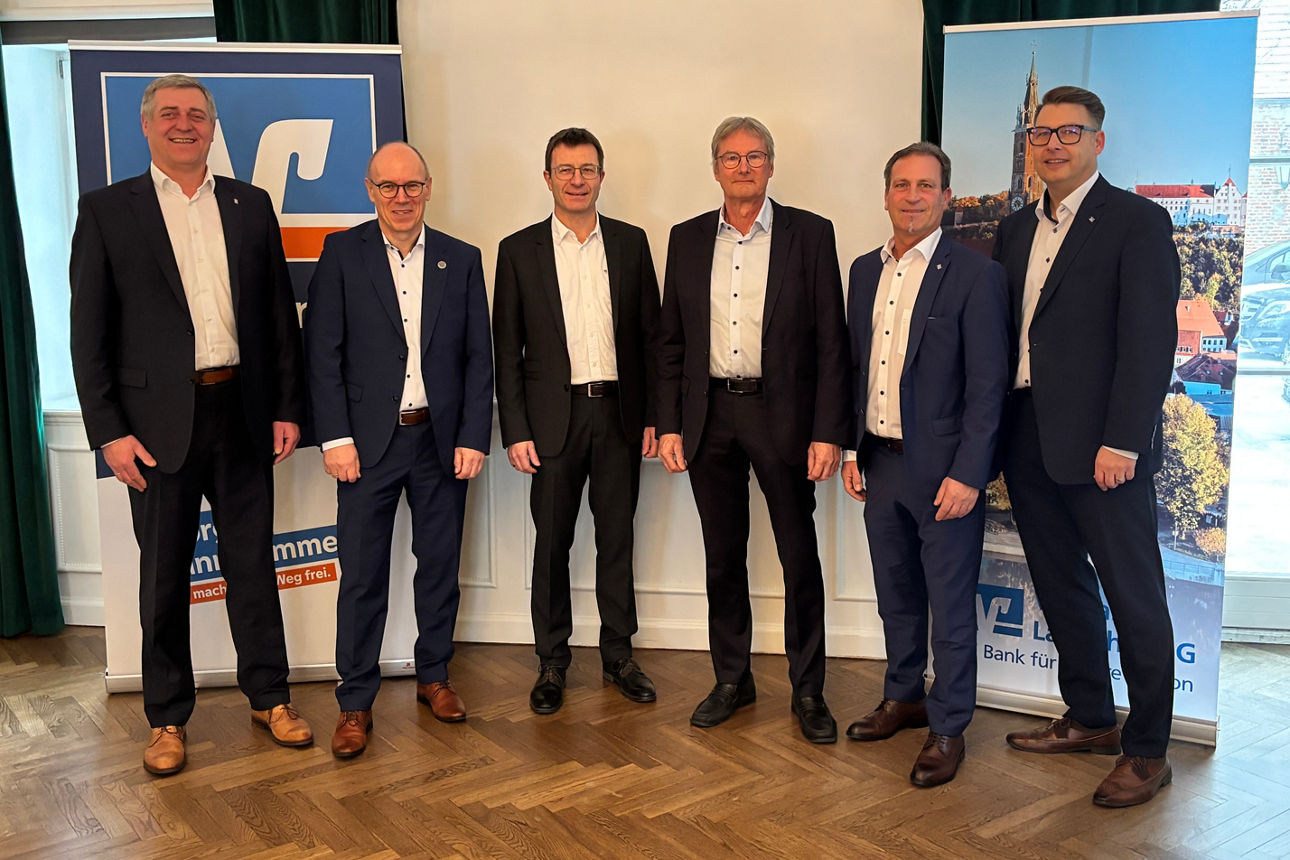 Sechs Männern in Anzügen lächeln in die Kamera. Sie stehen in einem großen Saal.