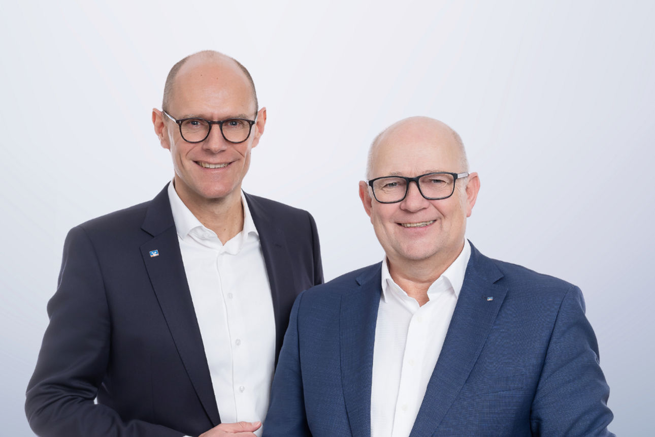 Beide Vorstände der Bank im Anzug, lächelnd. 